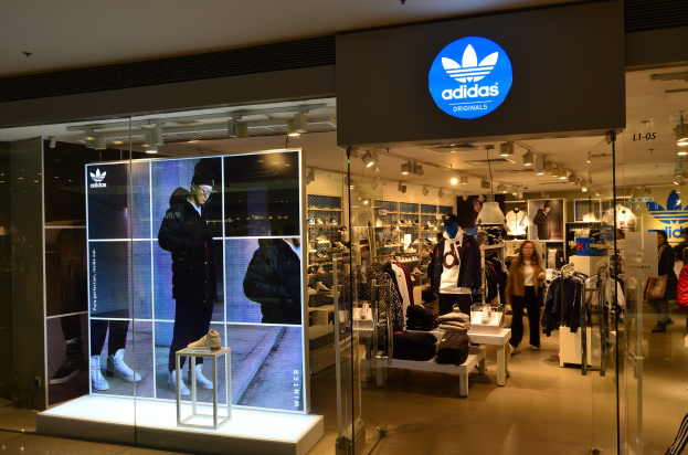 Außenansicht eines adidas Originals-Ladens in einem Einkaufszentrum mit Schaufensterpuppen, aufgehängten Kleidungsstücken, ausgestellten Schuhen und beleuchteter Markierung.