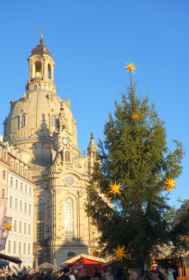 Ein geschäftiger Weihnachtsmarkt in Dresden, Deutschland, mit einem großen geschmückten Weihnachtsbaum vor einer Kirche, umgeben von vielen Menschen und einem Banner auf der linken Seite unter einem sichtbaren Himmel.