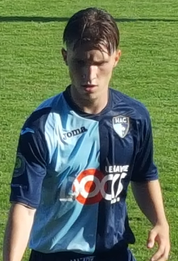 Ein junger Mann in einem blauen T-Shirt und schwarzen Shorts auf einem Fußballfeld während eines Spiels mit einem Ergebnis von 0:0.