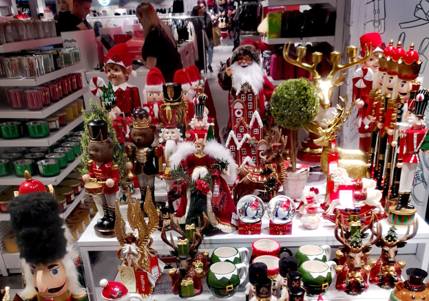 Ladeninneres weihnachtlich dekoriert mit Tischen, die Spielzeug, Tassen und andere Gegenstände zeigen, Regalen mit Waren und Menschen im Hintergrund bei festlichen Lichtern und einer Wand.