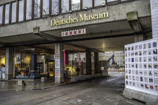 Außenansicht des Deutschen Museums in Berlin, das ein großes Glasgebäude mit Säulen, einem Namensschild und einer informativen Anzeige auf der rechten Seite sowie Bäumen und anderen Gebäuden im Hintergrund zeigt.