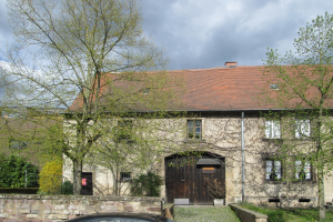 Ein Auto, das vor einem steinernen Gebäude mit einem Dach, Fenstern, einer Tür, einem Rohr, einer Wand, einigen Pflanzen, Gras, einer Gruppe von Bäumen und einem bewölkten Himmel geparkt ist.