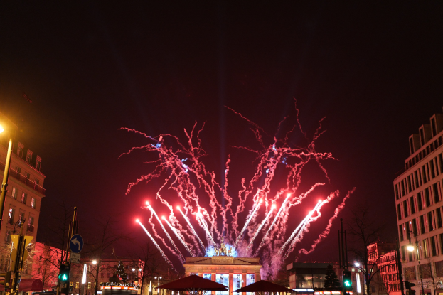 Eine belebte Straßen Szenerie in Berlin an Silvester, voller Menschen, Fahrzeuge und Gebäude, beleuchtet von Feuerwerk und Gebäudelichtern, die eine festliche Atmosphäre schaffen.