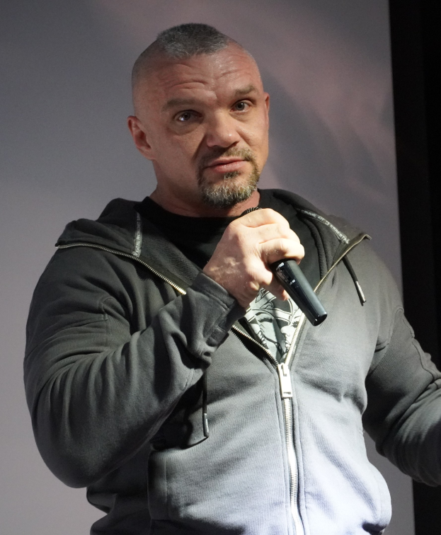 Ein Mann in einer schwarzen Kapuzenjacke und einem schwarzen Anzug mit einem weißen Hemd, glattem Haar und einem selbstbewussten Ausdruck hält ein Mikrofon, waßrend er vor einer Wand steht.