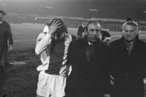 Schwarzes und weißes Bild einer Gruppe von Männern auf einem Fußballfeld mit einem Stadion im Hintergrund, die nach einem Sieg bedrückt aussehen.