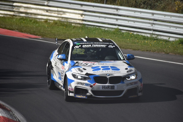 Ein BMW M4 DTM-Rennwagen mit Text und Zahlen auf der Karosserie, der auf einer Rennstrecke fährt, umgeben von Gras, einem Zaun und Pflanzen, wobei ein Rad sichtbar ist.