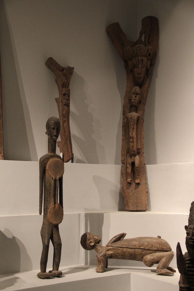 Eine Gruppe von Holzskulpturen auf einer weißen Fläche in einem Museum ausgestellt, mit einer Wand im Hintergrund.