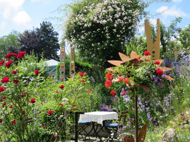 Ein farbenfroher Garten auf der Chelsea Flower Show mit einem zentralen Tisch mit einer Decke, umgeben von bunten Blumen und Pflanzen, mit Bäumen und einem bewölkten Himmel im Hintergrund.