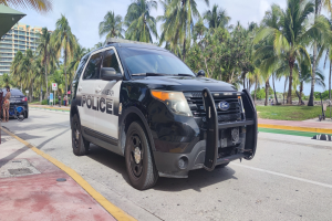 Polizeiauto am Straßenrand geparkt mit Menschen, Fahrrädern, Gras, Bäumen, Gebäuden und einem klaren blauen Himmel im Hintergrund.