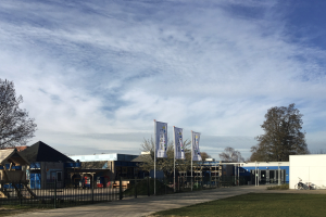Großes Gebäude mit einem Rasenbereich davor, umgeben von einem Metallzaun, mit einem Weg, Bäumen, Pfosten, Bannern, einem geparkten Fahrrad, nahegelegenen Häusern und einem bewölkten Himmel, als das neue Spielgelände der Schule identifiziert.