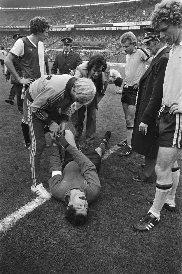 Schwarzes und weißes Foto eines Fußballspielers, der auf dem Boden liegt und von einer Gruppe Männer umgeben ist, mit einem Stadionpublikum im Hintergrund; der Spieler scheint verletzt zu sein.