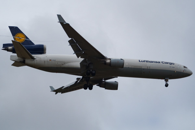 Ein Lufthansa Cargo Boeing 777-300ER im Flug, mit ausgebreiteten Flügeln und sichtbaren Triebwerken, mit der Aufschrift "Lufthansa Cargo" auf der Rumpfseite vor einem bewölkten Himmel.