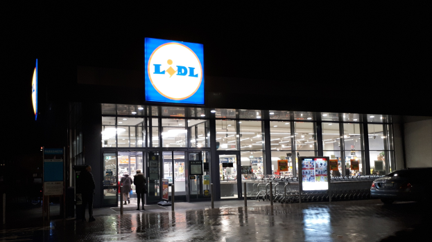 Ein Lidl-Laden bei Nacht mit Menschen davor, einem Auto rechts daneben und von der Decke beleuchtet.
