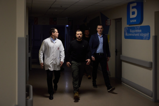 Eine Gruppe von Männern, die einen Krankenhausflur entlanggehen, mit Deckenbeleuchtung, einer Tür auf der rechten Seite und Texttafeln auf der linken Seite, die besorgt aussehen.