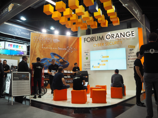Eine Gruppe von Menschen an einem IT-Cube Systems Forum Orange Cyber Security Stand auf einer Messe, mit Stühlen, einem Bildschirm, Regalen, Schildern, Deckenleuchten und einer Tafel mit Text.