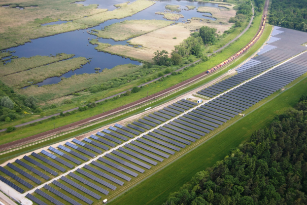 Luftaufnahme einer Solaranlage mit Panelen, einem Zug auf einer Schiene, umgeben von Bäumen, Gras, Wasser und Feldern.
