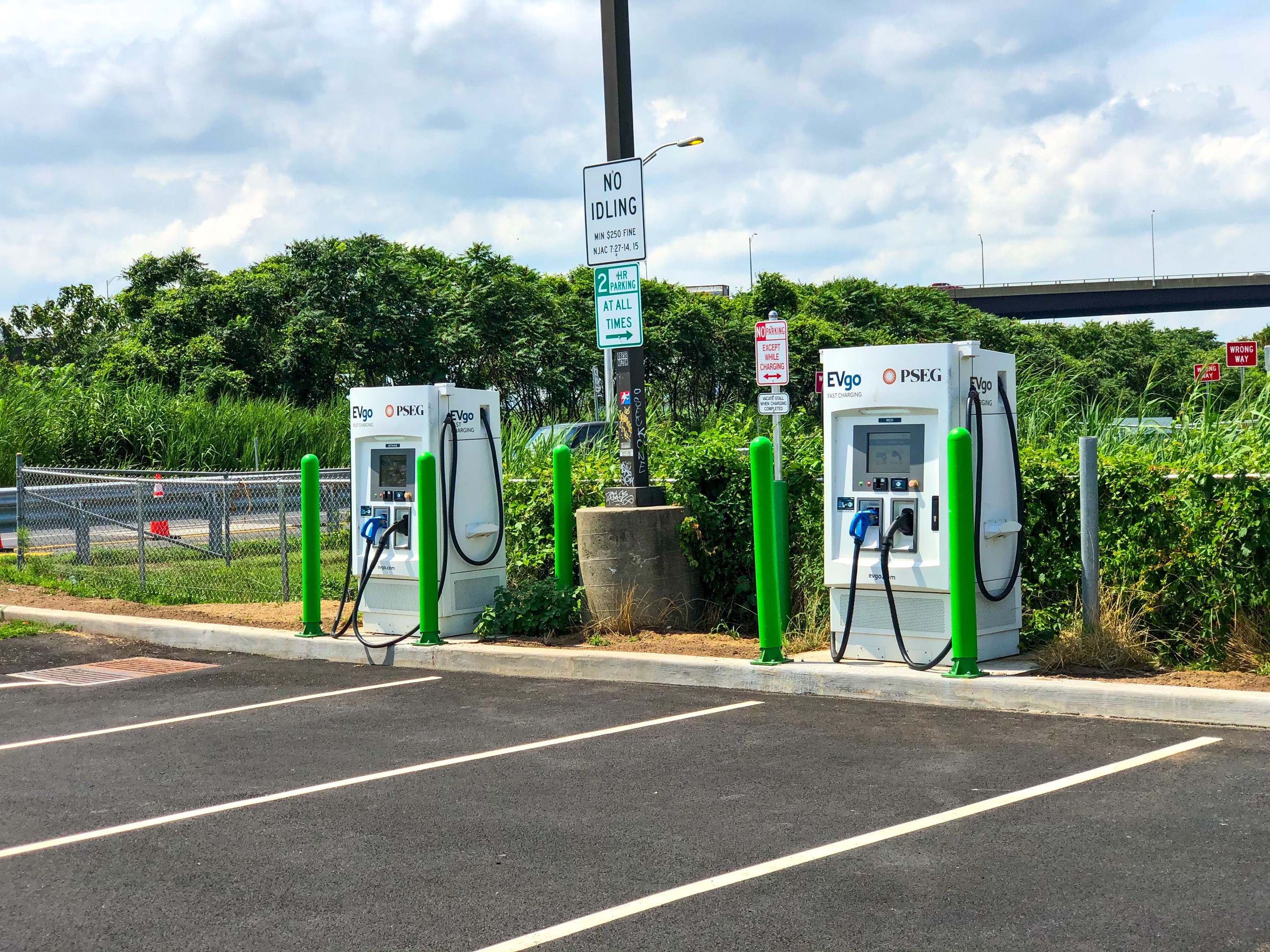 Elektrische Ladestation für Fahrzeuge auf einem Parkplatz mit umgebenen Infrastruktur, Grünflächen und einer Brücke im Hintergrund bei bewölktem Himmel.