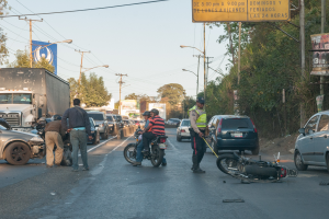 Eine Gruppe von Menschen steht um ein verunglücktes Motorrad am Straßenrand mit mehreren Fahrzeugen, darunter ein Lastwagen, und einer Hintergrundlandschaft aus Bäumen, Pfählen, Laternen, Schildern und Himmel.