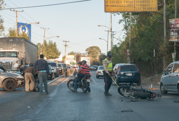 Eine Gruppe von Menschen steht um ein verunglücktes Motorrad am Straßenrand mit mehreren Fahrzeugen, darunter ein Lastwagen, und einer Hintergrundlandschaft aus Bäumen, Pfählen, Laternen, Schildern und Himmel.