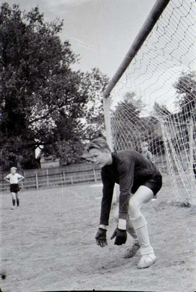 Schwarz-weiß-Foto eines jungen Jungen, der auf einem umzäunten Feld mit Bäumen und einem klaren Himmel Fußball spielt, mit einer Person im Vordergrund und anderen Kindern im Hintergrund.
