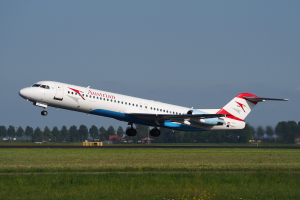 Ein Embraer ERJ-190-Flugzeug der Austrian Airlines beim Start am Frankfurter Flughafen, mit grünem Gras am Boden und Bäumen und Gebäuden im Hintergrund, unter einem klaren Himmel.