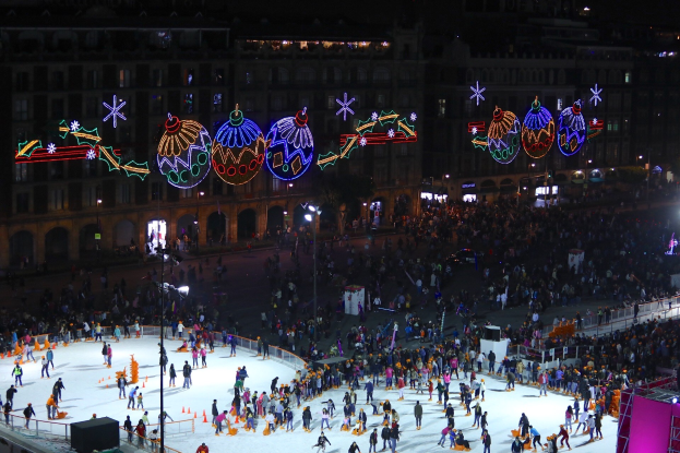 Große Menschenmenge auf einer Eisbahn mit festlichen Dekorationen, umgeben von Geländern und Pfählen, unter Straßenlaternen vor dunklem Hintergrund.