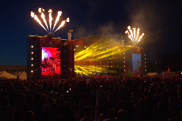 Menschenmenge auf einem Musikfestival mit Performern auf einer hell erleuchteten Bühne, umgeben von Bannern, Zelten links und Feuerwerk am dunklen Himmel.