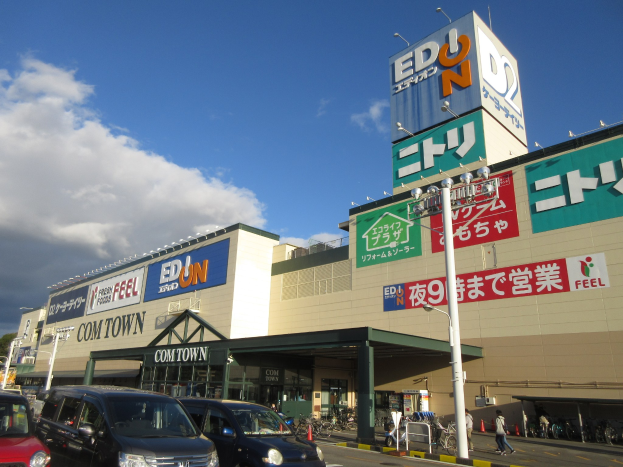 Ein Einkaufszentrum mit parkenden Autos, Fußgängern auf dem Gehweg, fahrenden Fahrzeugen, Fahrrädern, Bäumen, Straßenmöblierung und Gebäuden unter einem bewölkten Himmel.