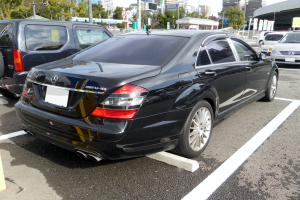 Ein schwarzer Mercedes S-Klasse in einer Parklücke mit anderen Autos, Gebäuden, Bäumen, Polen, Plakatwänden und einem klaren blauen Himmel geparkt.