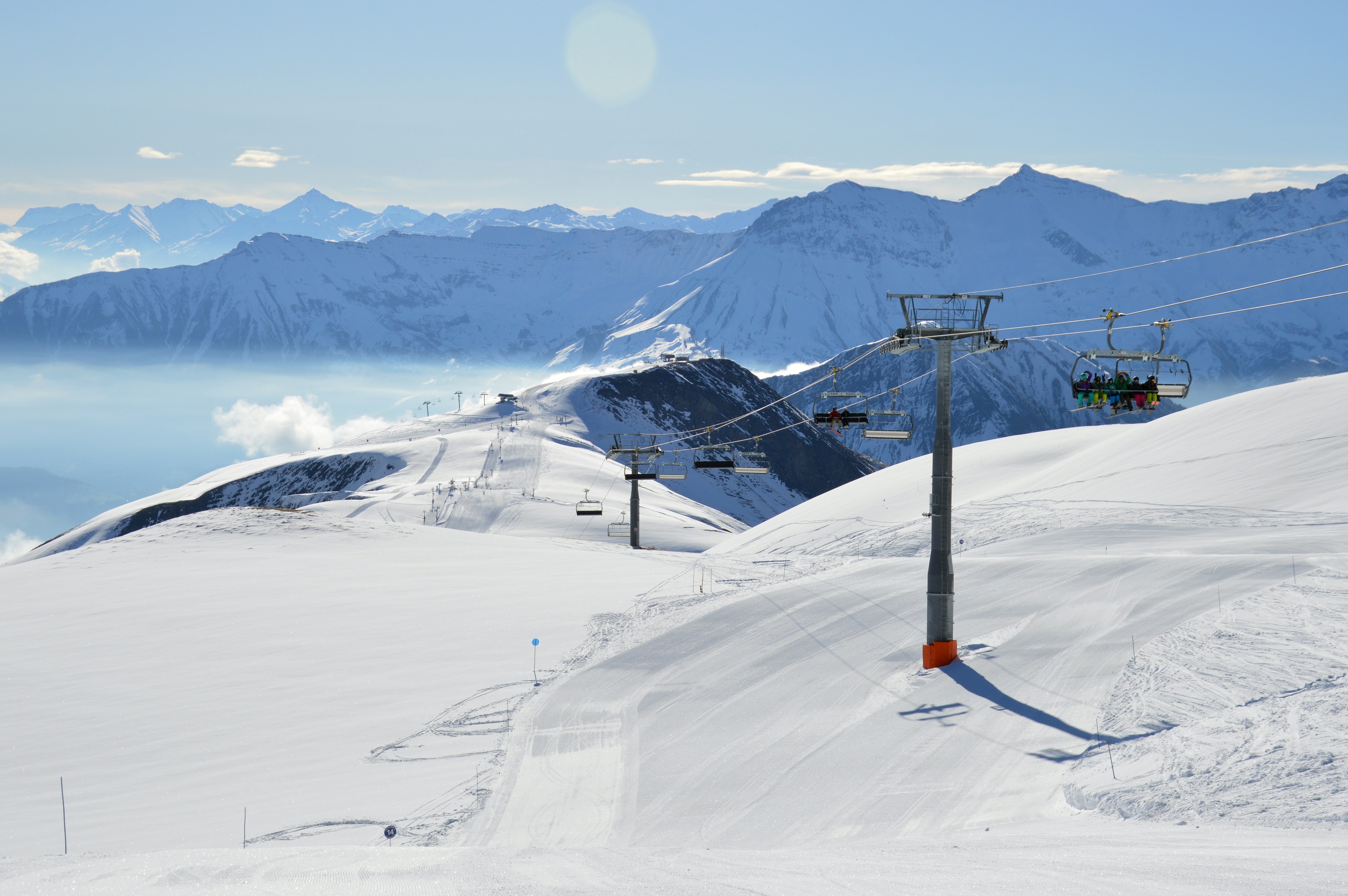 Skilift fährt eine schneebedeckte Bergflanke hoch, mit Passagieren, die von Stützen und Seilen unterstützt werden, vor einem Hintergrund aus schneebedeckten Hügeln und bewölktem Himmel.