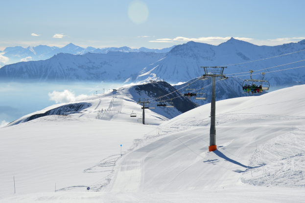 Skilift fährt eine schneebedeckte Bergflanke hoch, mit Passagieren, die von Stützen und Seilen unterstützt werden, vor einem Hintergrund aus schneebedeckten Hügeln und bewölktem Himmel.