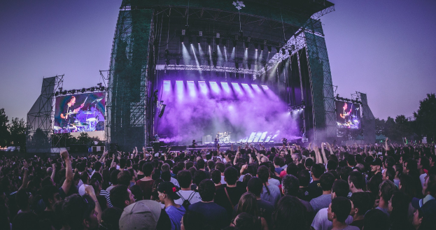 Eine große Menschenmenge auf einem Musikfestival schaut auf einer beleuchteten Bühne mit Instrumenten und Bildschirmen, die vor einem Gebäude, Bäumen und einem klaren Himmel aufgebaut ist.
