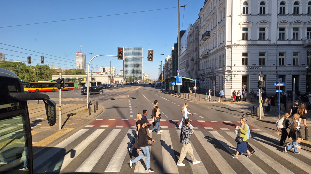Gruppe von Menschen, die eine Straße an einer Ampel überqueren, mit einem Bus auf der linken Seite, Fahrzeugen auf der Straße, Verkehrszeichen, Laternenpfähle, Gebäude, Bäume und einen klaren blauen Himmel.