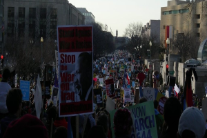 Eine Gruppe von Menschen, die auf einer Straße protestieren und Plakate halten, mit Gebäuden, Bäumen, Laternenmasten und dem Himmel im Hintergrund.