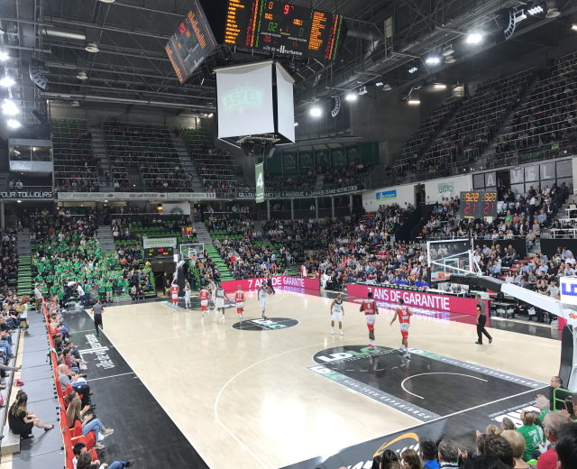 Basketballspiel in einer großen Arena mit Zuschauern, mit Beteiligung der Mannschaften Asvel und Galatasaray, unter Stadionbeleuchtung und einem Scoreboard.