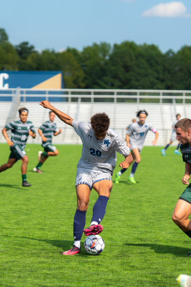 Eine Gruppe junger Männer, die Fußball auf einem Feld mit Bäumen und einem klaren blauen Himmel spielen, ein Spieler in Aktion mit Turnschuhen.