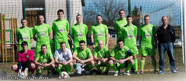 Gruppe von Männern in grünen T-Shirts auf einem Fußballfeld mit einem Ball und einem Torpfosten, Bäume und Gebäude im Hintergrund.