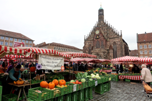 Ein belebter Markt in Nürnberg, Deutschland, mit farbenfrohen Obst- und Gemüsesorten auf dem Markt, Menschen mit Taschen in der Hand und Zelten um den Markt herum, mit Gebäuden und einem Uhrenturm im Hintergrund unter einem sichtbaren Himmel.