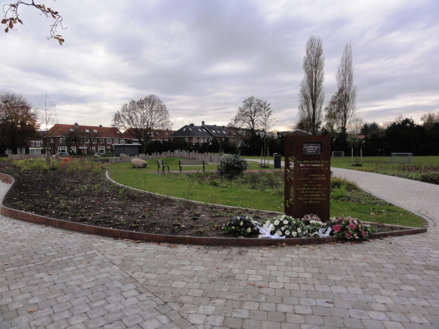 Eine Gedenkstätte im Park mit einem Backsteinweg, umgeben von Grün, Bänken und Bäumen, mit Gebäuden und einem bewölkten Himmel im Hintergrund und einer Tafel mit Text auf der rechten Seite.