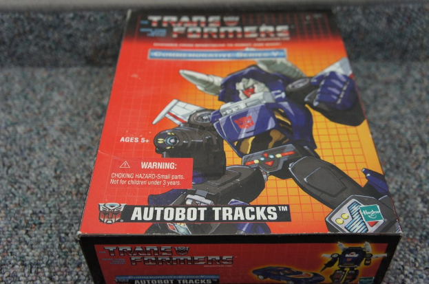 Ein roter, oranger und blauer AUTOBOT TRACKS-Roboterkasten mit "AUTOBOT TRACKS"-Text oben auf einem unscharfen schwarzen Hintergrund.