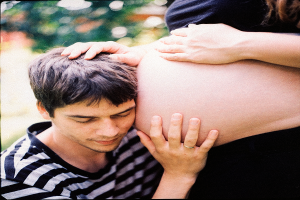 Ein Mann und eine Frau stehen nebeneinander, wobei der Mann zärtlich den Bauch der Frau berührt und sich auf ihr ungeborenes Kind konzentriert.