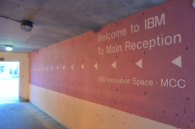 Eine Wand mit den Worten "Willkommen bei IBM", beleuchtet von Deckenlampen, mit einer Tür im Hintergrund, die "IBM Innovationsraum MCC" markiert.