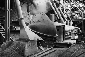 Ein Schwarz-Weiß-Foto einer großen Maschine in einer Fabrik, umgeben von verschiedenen Maschinen, Strukturen und Eisenbahnschienen unten.