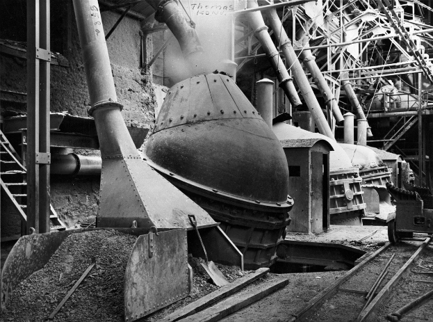 Ein Schwarz-Weiß-Foto einer großen Maschine in einer Fabrik, umgeben von verschiedenen Maschinen, Strukturen und Eisenbahnschienen unten.