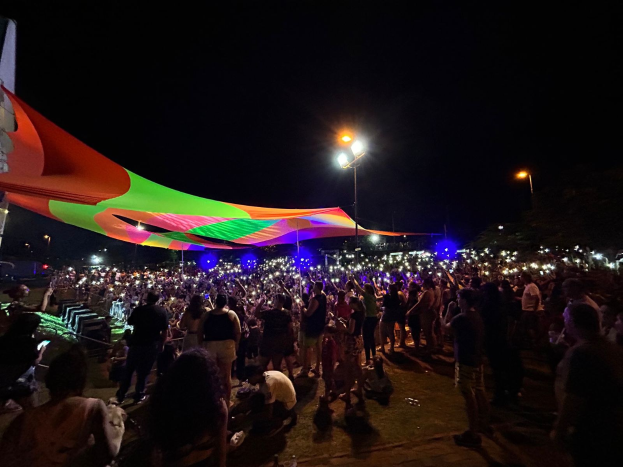 Eine große Menschenmenge versammelt sich unter bunten Lichtern nachts vor einem Zelt, mit einem Gebäude auf der linken Seite und dem Nachthimmel im Hintergrund, das das 2018er Kunstfest zeigt.