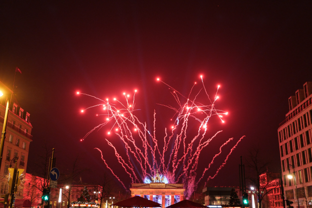 Eine Stadtstraße in Berlin an Silvester, gefüllt mit Gebäuden, Bäumen, Laternenmasten, Verkehrszeichen, Zelten und Menschen, mit einem von Feuerwerk erhellten Himmel im Hintergrund.