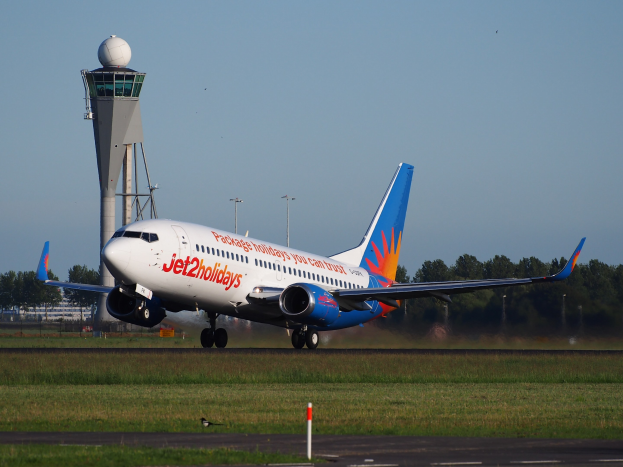 Ein Jet2holidays Airbus A320-200 startet von Frankfurt Airport mit grünem Gras darunter, einem hohen Turm im Hintergrund, Bäumen und Gebäuden in der Ferne und einem hellblauen Himmel mit Sonnenlicht, das das aufsteigende Flugzeug erhellt.