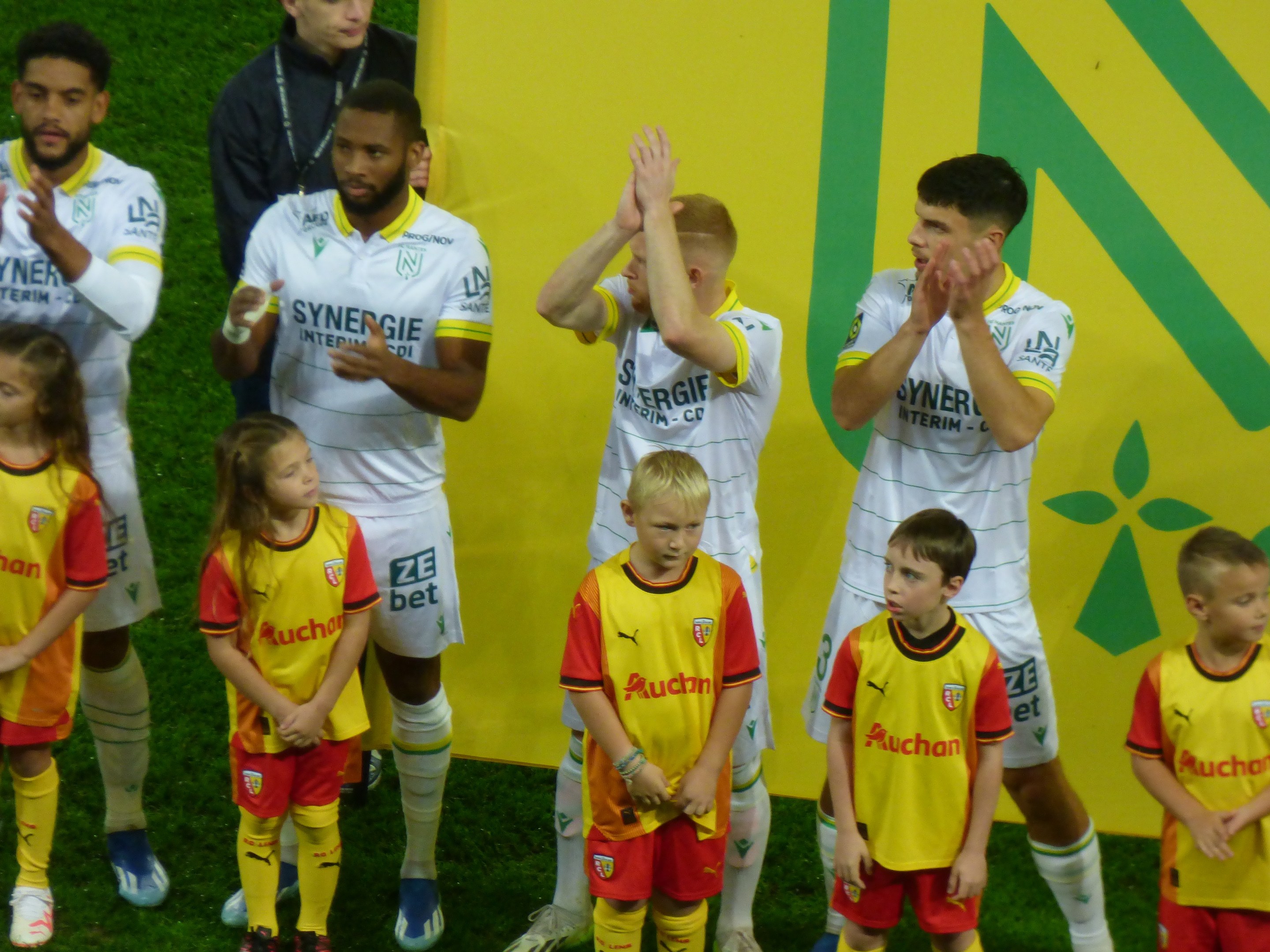 Eine Gruppe von Fußballspielern in Uniformen klatscht auf einem Ligue-1-Feld in Feierlaune, mit einem "Ligue-1"-Schild im Hintergrund.