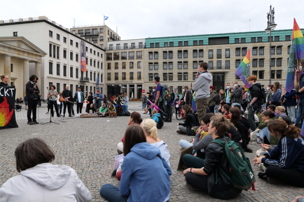 Eine Gruppe von Menschen sitzt auf dem Boden vor einer Menge mit Fahnen und Spruchbändern, mit einer Person, die in ein Mikrofon spricht, einer Statue und Gebäuden im Hintergrund während einer anti-schwulen Demonstration in Berlin.