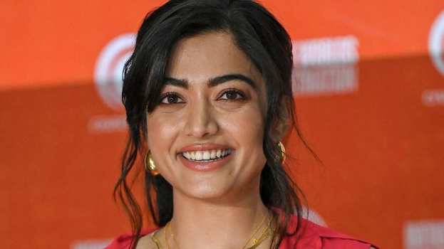 Eine Frau in einem roten Kleid mit einem eleganten Dutt und dezentem Make-up steht strahlend da, trägt schwarze High Heels und hat ein hellgelbes Banner mit der Aufschrift "Sonam Kapoors Schönste Looks vom Cannes Film Festival" im Hintergrund.